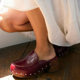 A close-up of a person wearing burgundy leather clog-style mule heels with small silver studs and wooden block heels, seated on a wooden floor with a white dress visible.