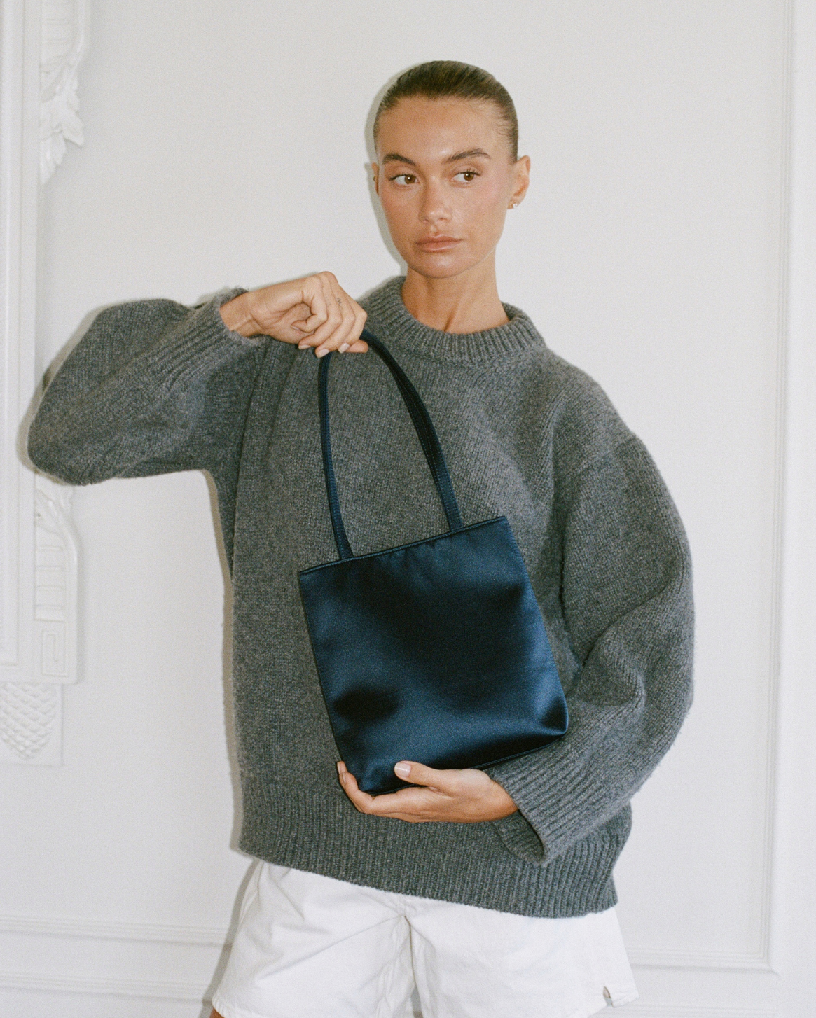 A model wearing a gray knit sweater and white shorts holds a dark navy satin tote bag against a white paneled wall.
