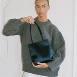 A model wearing a gray knit sweater and white shorts holds a dark navy satin tote bag against a white paneled wall.