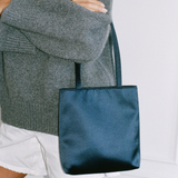 A model wearing a gray knit sweater and white shorts holds a dark navy satin tote bag against a white paneled wall.
