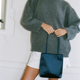 A person wearing an oversized heathered gray knit sweater and white shorts, holding a dark navy satin tote bag against a white interior wall.