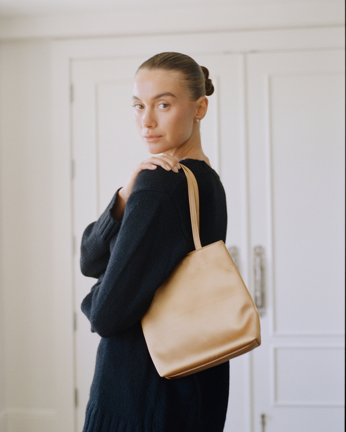 A woman wearing a dark navy knit sweater carries a large, structured tan leather tote bag over her shoulder, standing in front of white paneled doors.