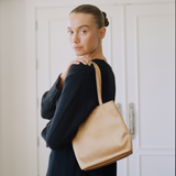 A woman wearing a dark navy knit sweater carries a large, structured tan leather tote bag over her shoulder, standing in front of white paneled doors.
