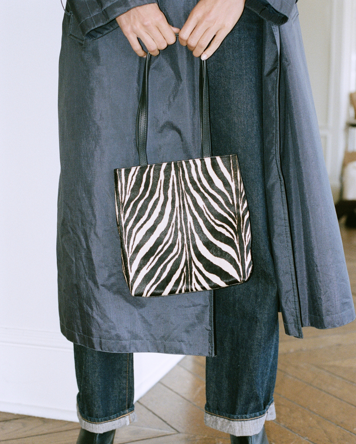 A person wearing a dark gray coat and blue jeans holds a black and white zebra-print tote bag in an indoor setting.
