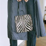 A person wearing a dark gray coat and blue jeans holds a black and white zebra-print tote bag in an indoor setting.