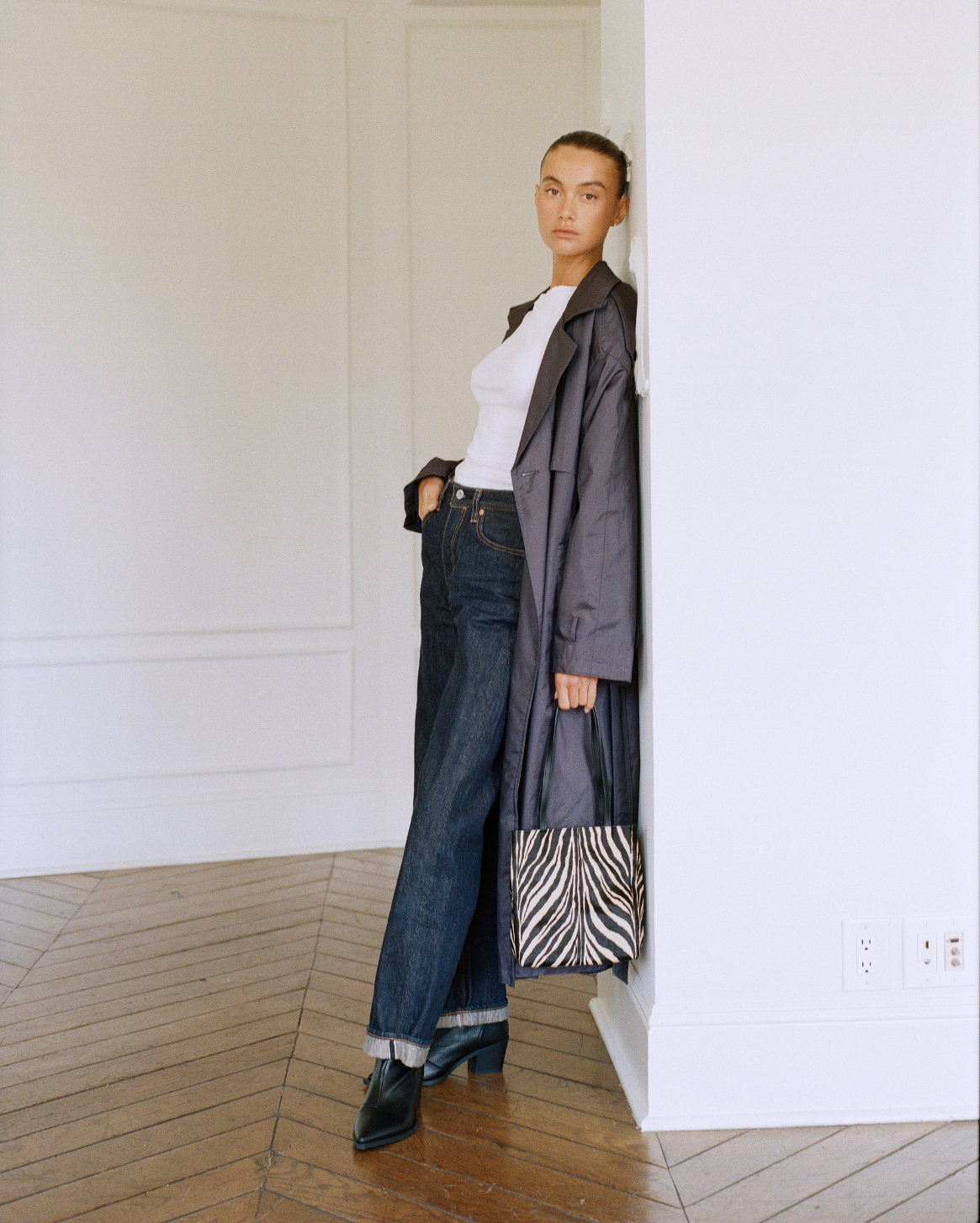 A model stands in a minimalist white room with wood floors, wearing a dark gray trench coat over a white top, dark blue jeans, and black shoes, while holding a black and white zebra-patterned tote bag.