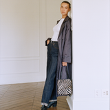 A model stands in a minimalist white room with wood floors, wearing a dark gray trench coat over a white top, dark blue jeans, and black shoes, while holding a black and white zebra-patterned tote bag.