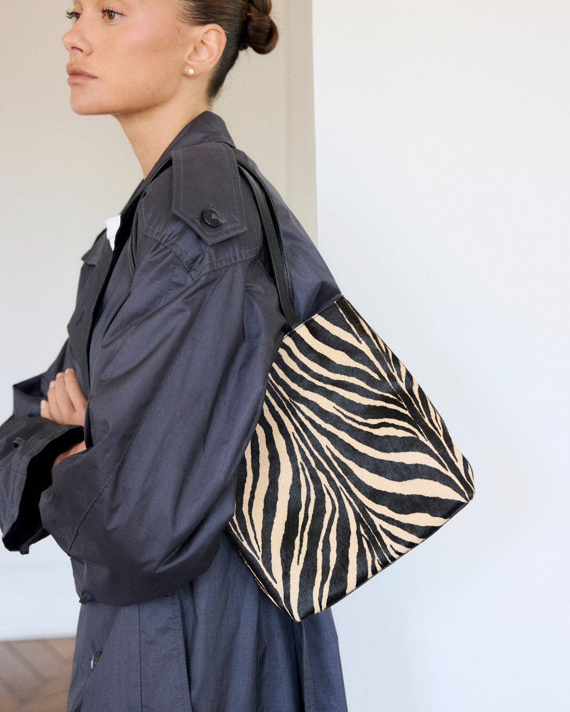 A model wearing a dark gray coat holds a black and white zebra-print tote bag in an indoor setting.