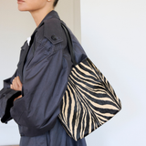 A model wearing a dark gray coat holds a black and white zebra-print tote bag in an indoor setting.
