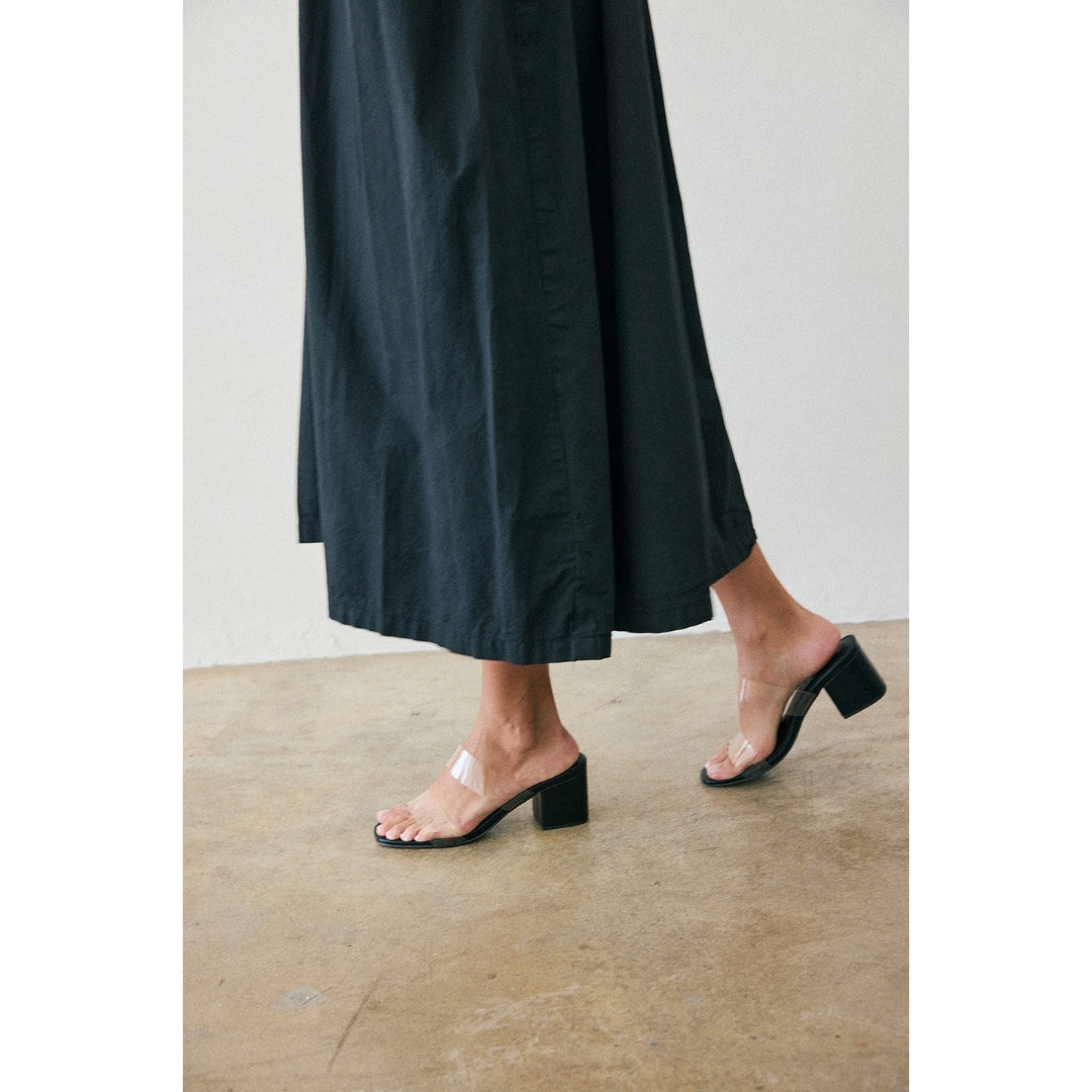 Person wearing a black dress and black heeled sandals with clear straps on a concrete floor.