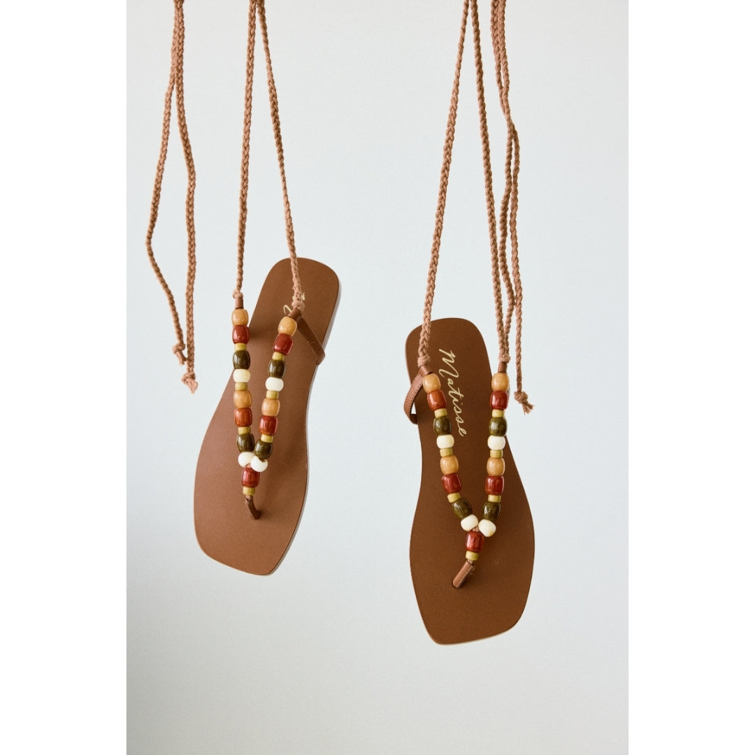 Pair of brown sandals with beaded straps hanging against a light gray background.