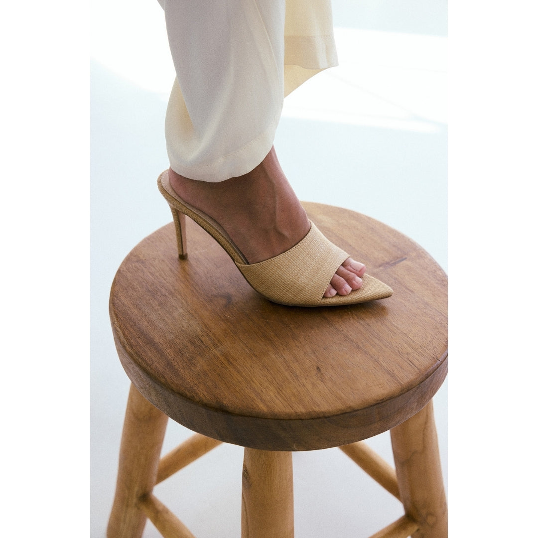 Beige high-heeled sandal on a wooden stool with a blurred background.