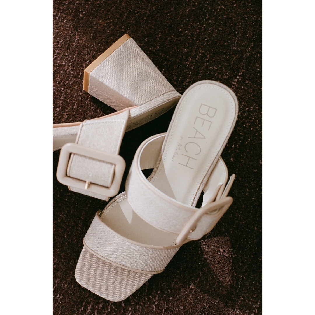 Pair of beige heeled sandals with a buckle on a brown textured surface.