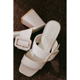 Pair of beige heeled sandals with a buckle on a brown textured surface.