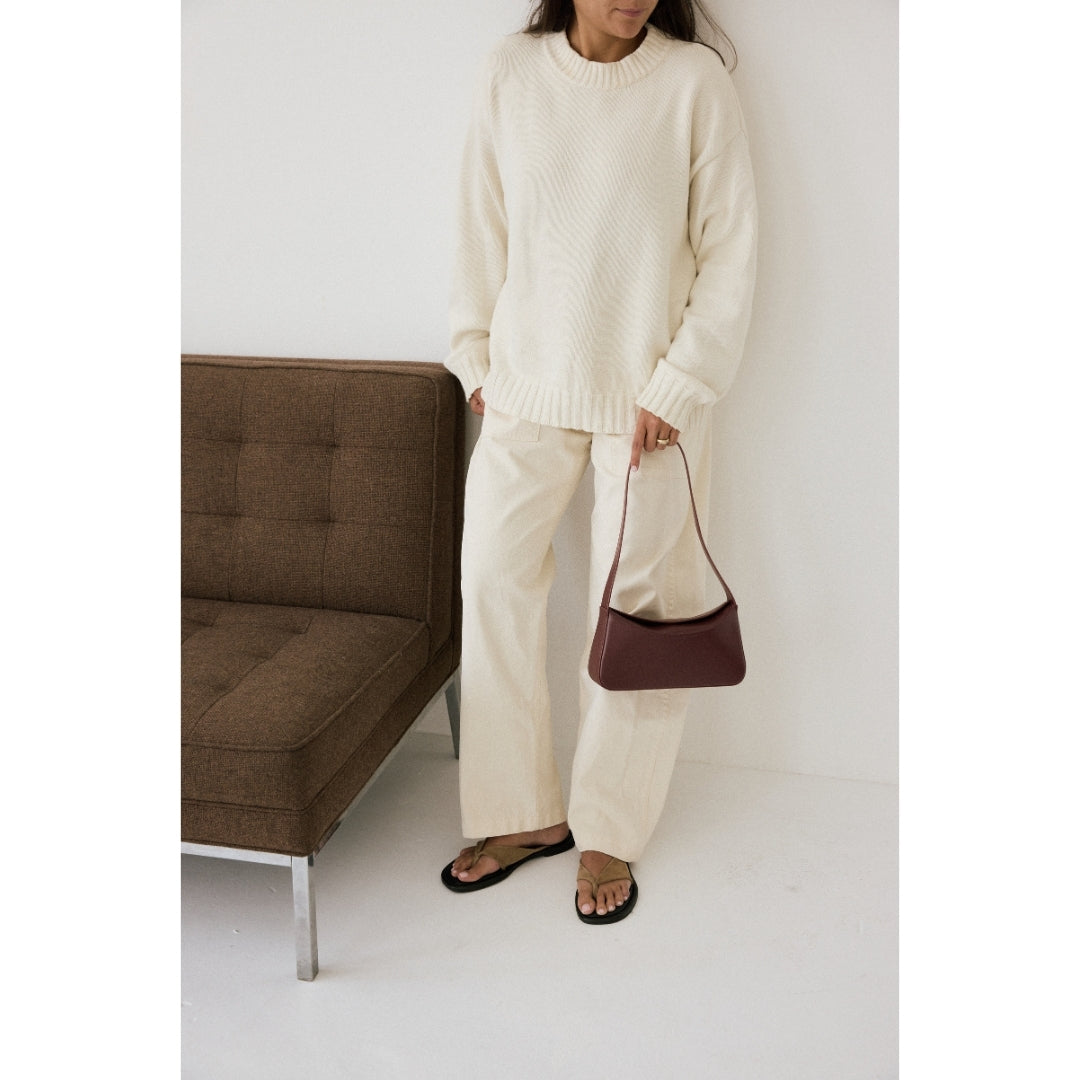 Person wearing a cream sweater and pants holding a brown handbag next to a brown sofa.