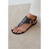 Black sandal with studded details on a foot against a neutral background.
