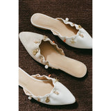 Pair of white and beige embellished mules on a brown textured surface.