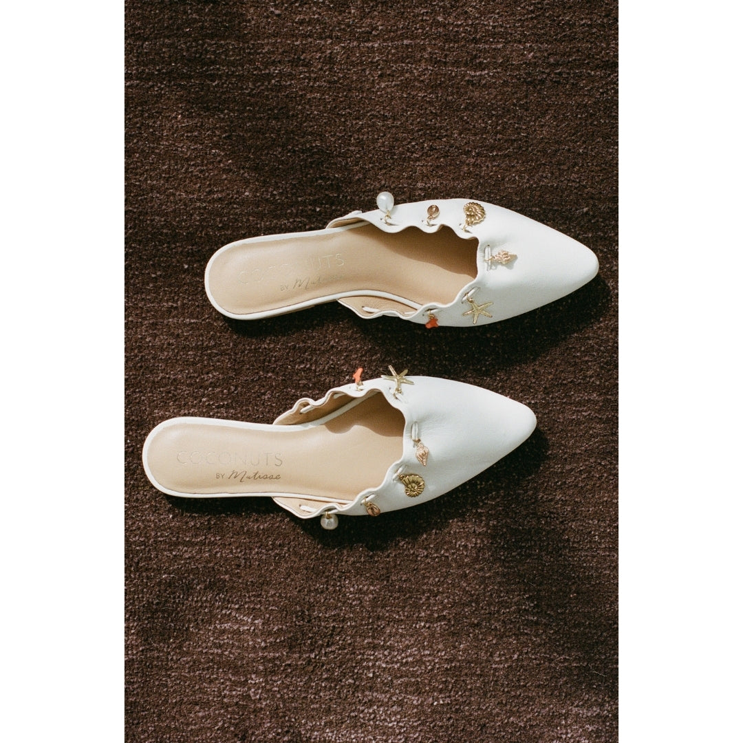 Pair of white pointed-toe mules with decorative elements on a brown textured surface.
