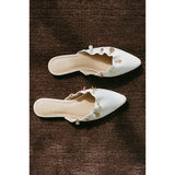 Pair of white pointed-toe mules with decorative elements on a brown textured surface.