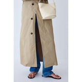 Person wearing a beige trench coat and blue jeans, holding a beige handbag on a light gray background.