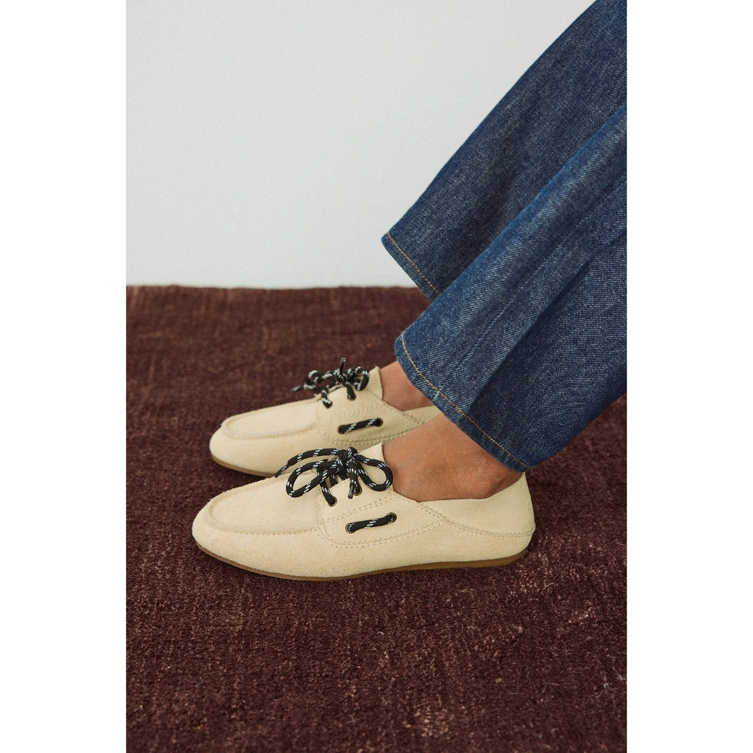Beige boat shoes with black laces worn with blue jeans on a brown carpet.