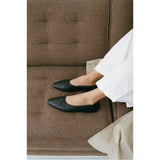 Person wearing black woven flatssitting on a brown upholstered chair.