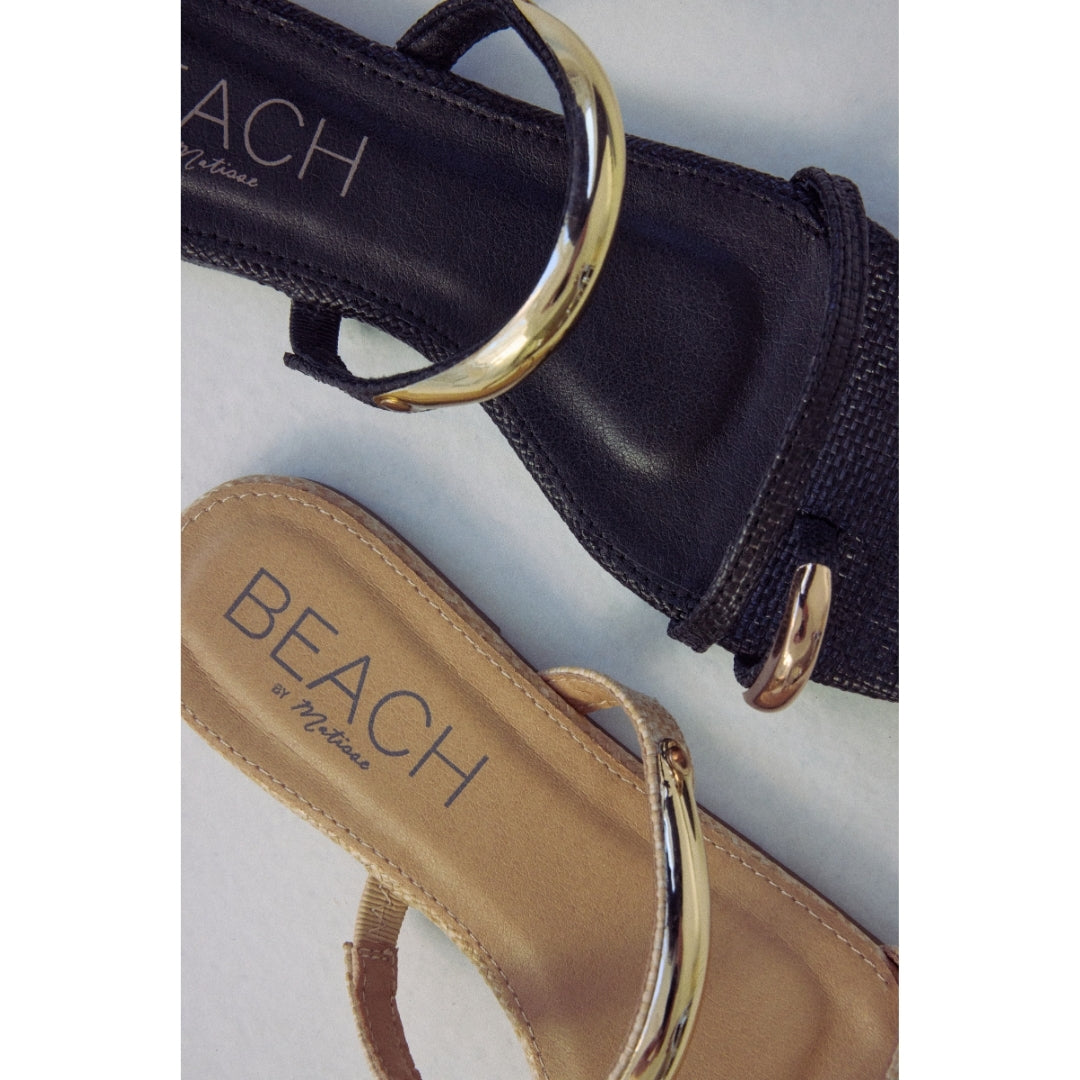 Close-up of black and beige sandals with gold straps and 'Beach by Matisse' branding on a light gray background.