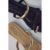 Close-up of black and beige sandals with gold straps and 'Beach by Matisse' branding on a light gray background.