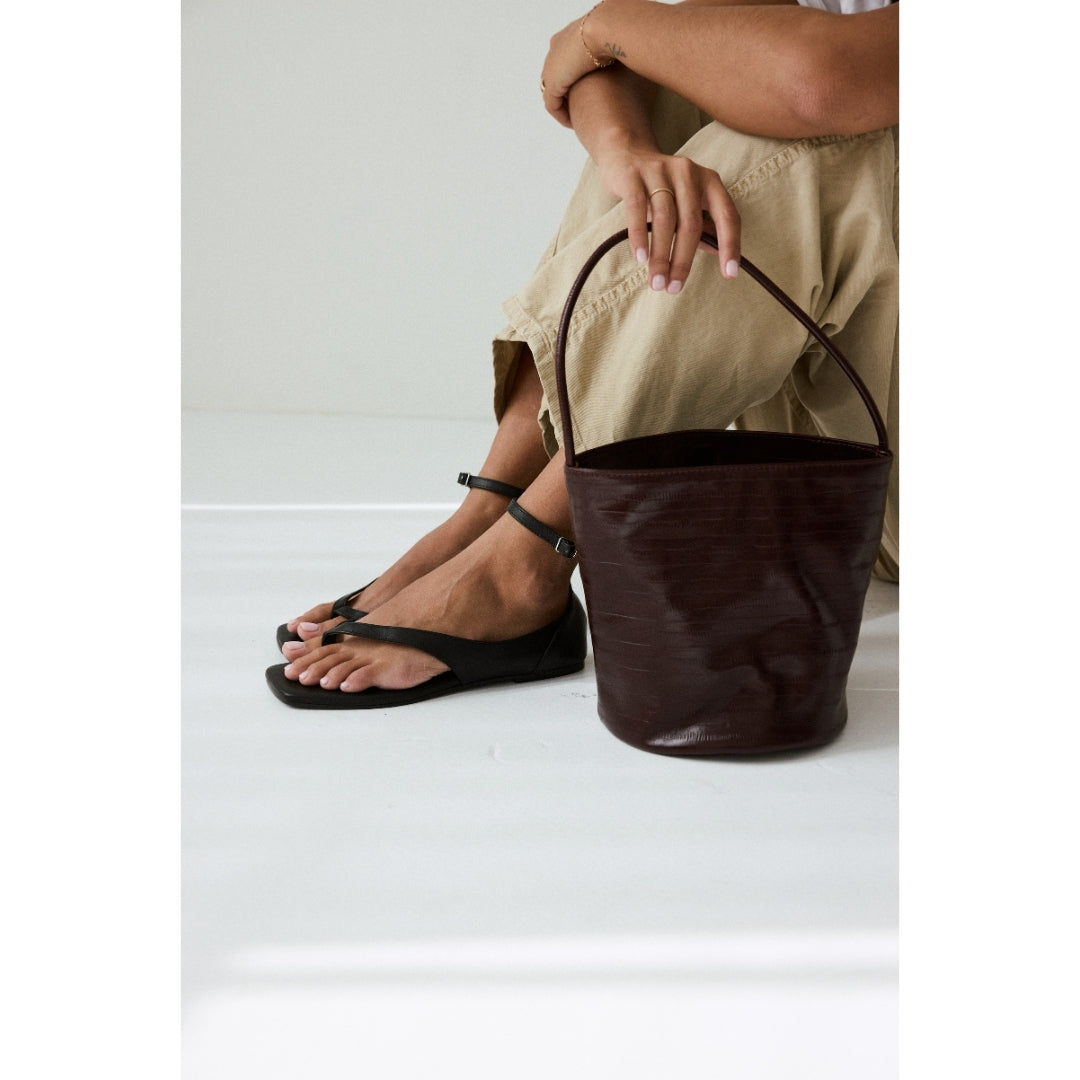 Person wearing black sandals and holding a burgundy leather bucket on a white background.
