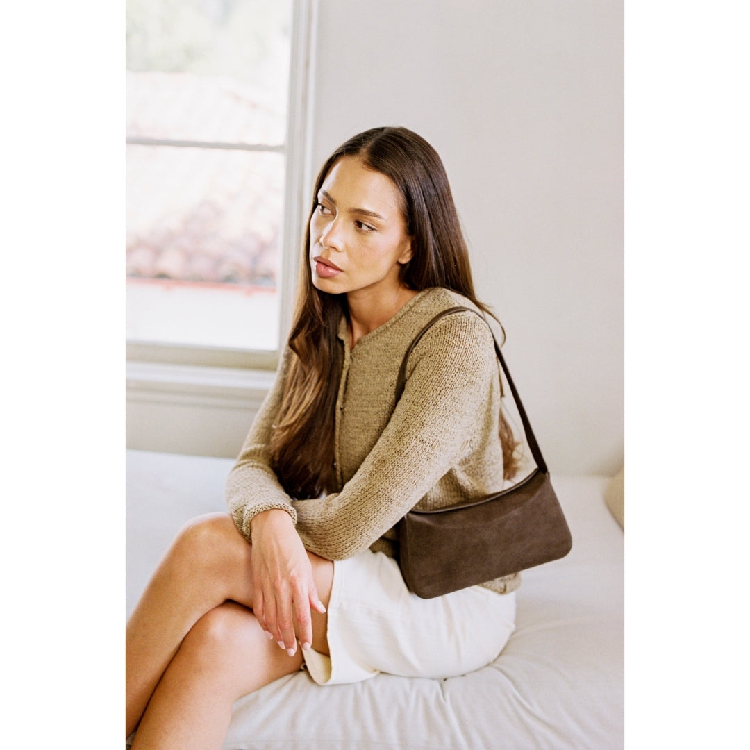 Woman sitting on a bed wearing a brown sweater and holding a brown handbag.