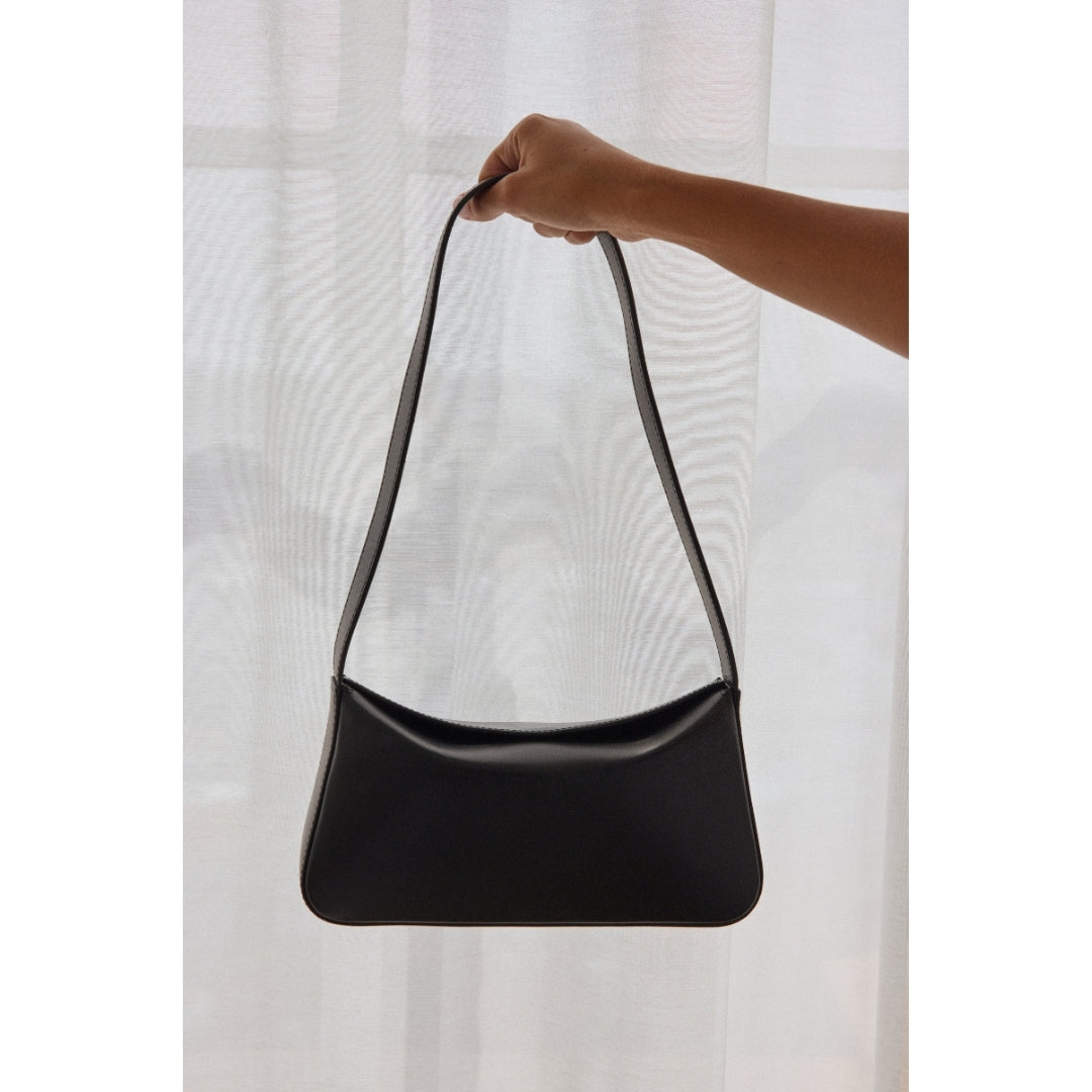 Black handbag held by a hand against a white curtain background.