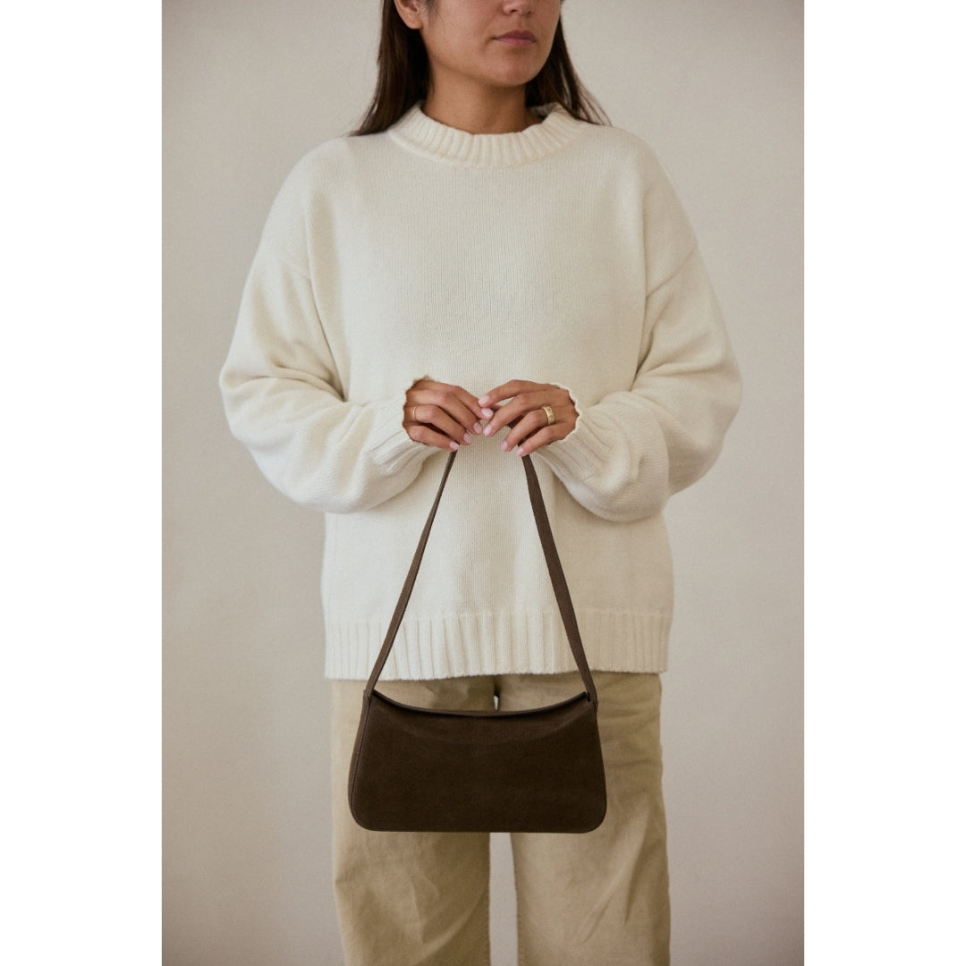 Person wearing a white sweater and holding a brown handbag against a neutral background.