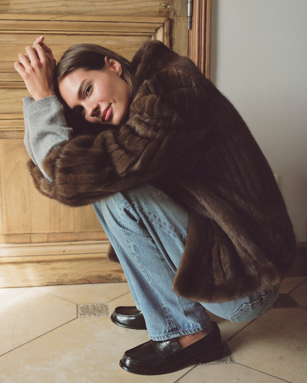 A woman crouches indoors wearing a voluminous dark brown fur coat over a light gray sweater, light blue jeans, and black loafers, posed against a wooden door and tiled floor.