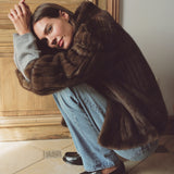 A woman crouches indoors wearing a voluminous dark brown fur coat over a light gray sweater, light blue jeans, and black loafers, posed against a wooden door and tiled floor.