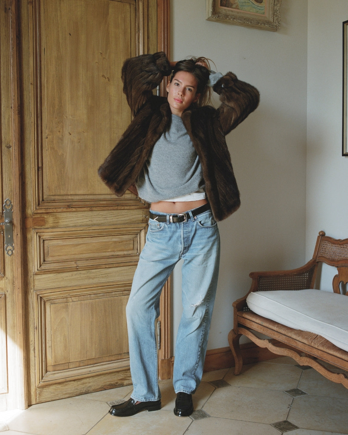 Person wearing a fur coat, gray sweater, and blue jeans in a room with wooden door and chair.
