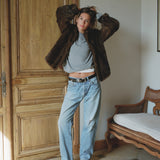 Person wearing a fur coat, gray sweater, and blue jeans in a room with wooden door and chair.