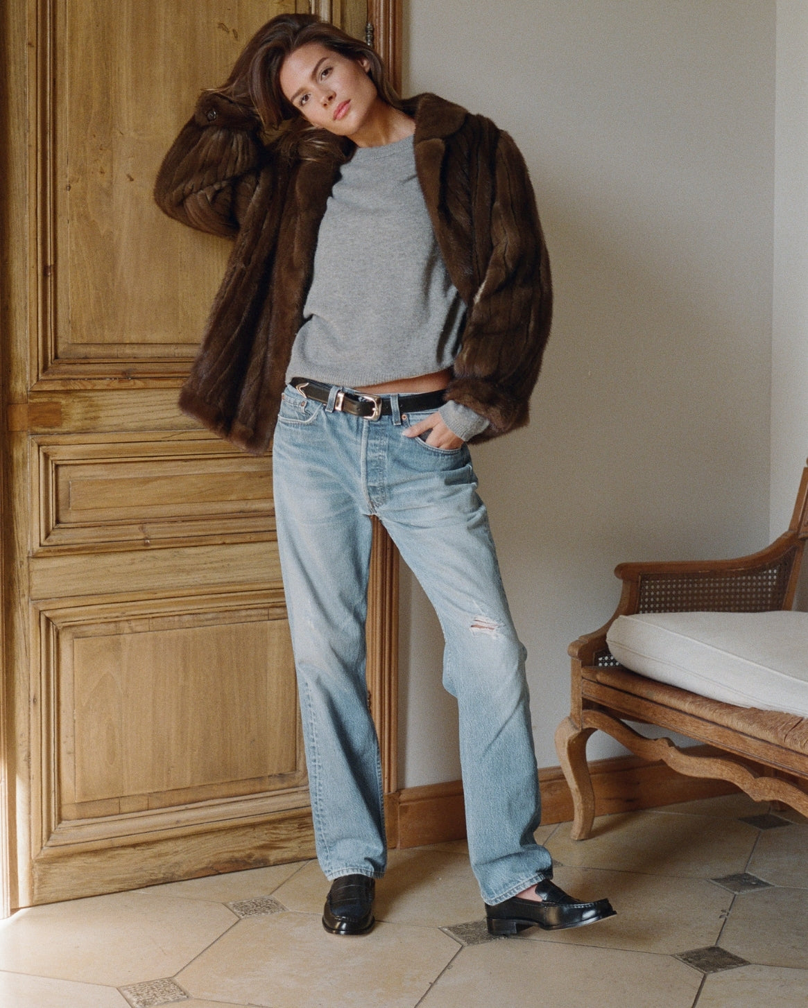 Woman wearing a brown fur coat, gray sweater, and blue jeans standing in a room with wooden paneling.