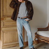 Woman wearing a brown fur coat, gray sweater, and blue jeans standing in a room with wooden paneling.