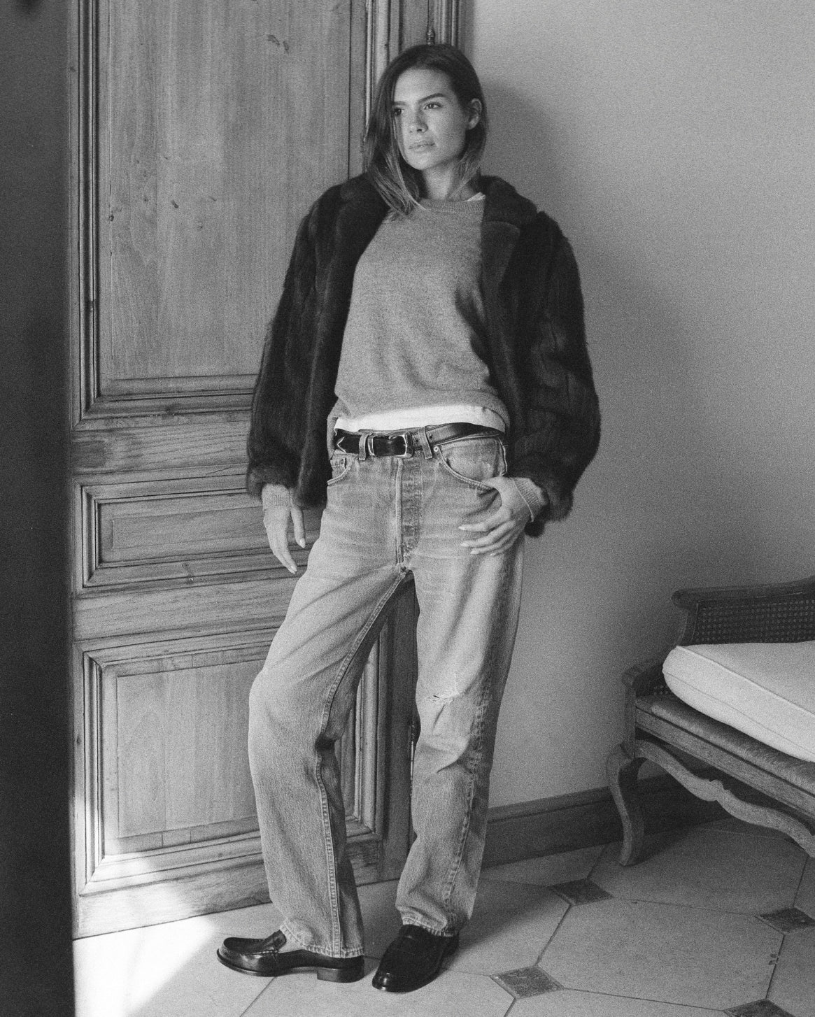 Person wearing a jacket, sweater, and jeans standing in a room with wooden paneling.