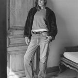 Person wearing a jacket, sweater, and jeans standing in a room with wooden paneling.