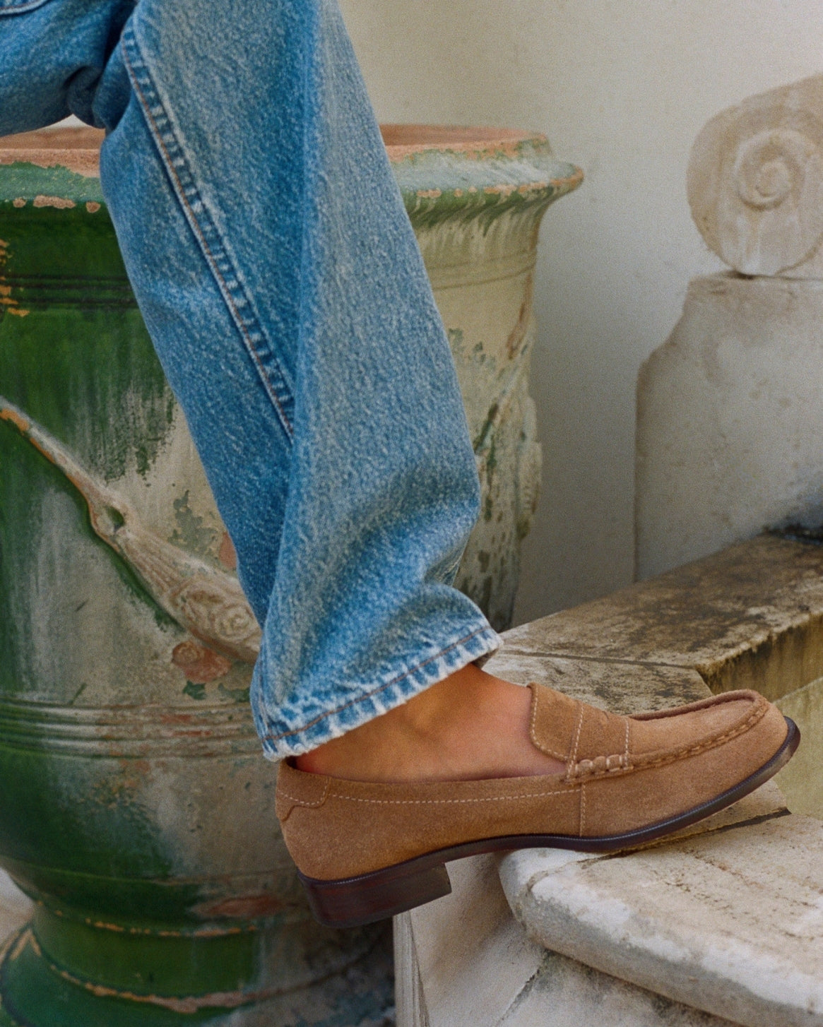 A person wearing blue jeans and a tan suede loafer sits on stone steps beside a large green planter.