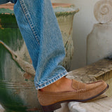 A person wearing blue jeans and a tan suede loafer sits on stone steps beside a large green planter.