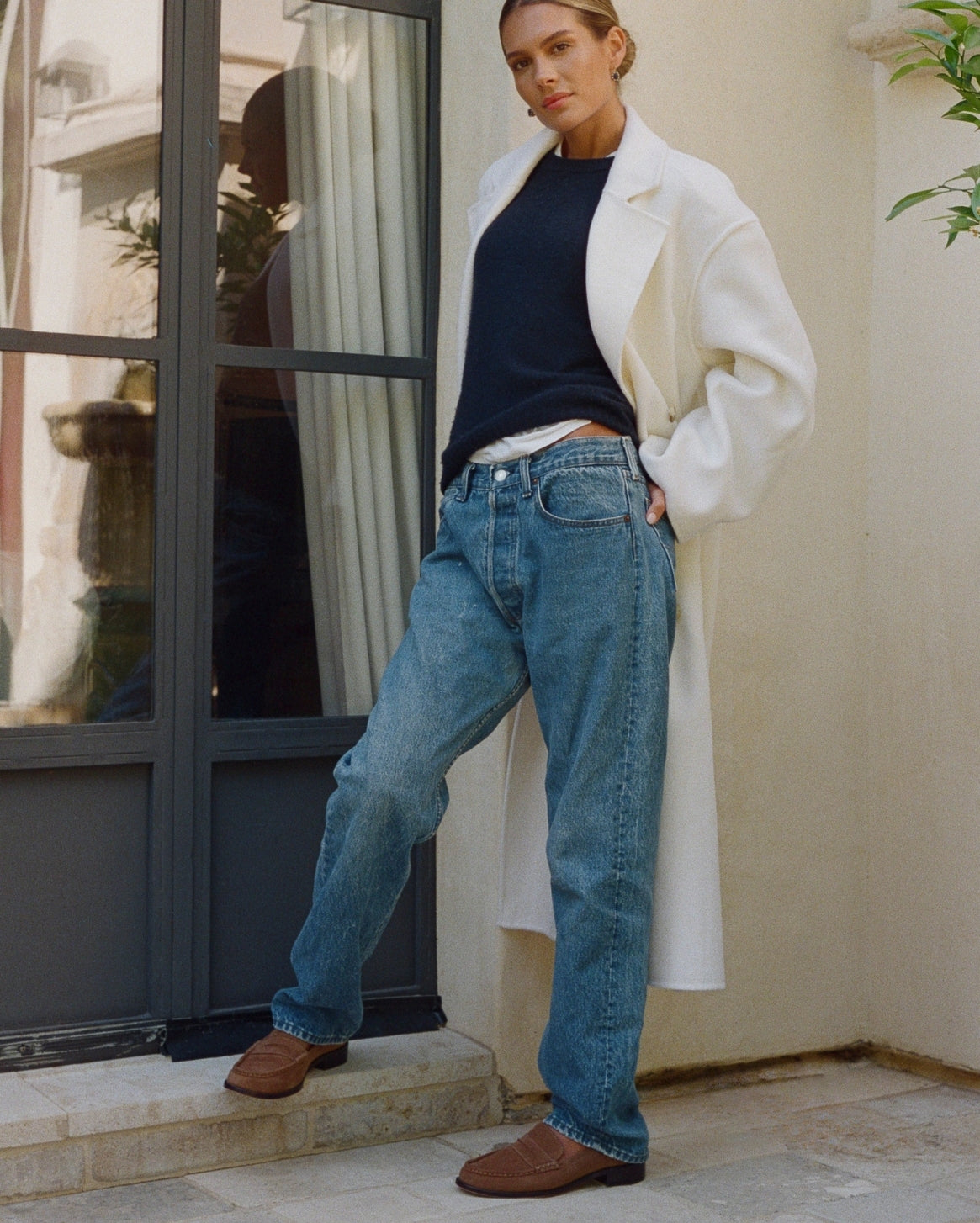 A woman models a pair of relaxed-fit blue jeans with a raw hem, styled with a white coat, navy top, and brown loafers, standing on stone steps in front of a beige wall and dark-framed glass door.
