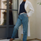 A woman models a pair of relaxed-fit blue jeans with a raw hem, styled with a white coat, navy top, and brown loafers, standing on stone steps in front of a beige wall and dark-framed glass door.