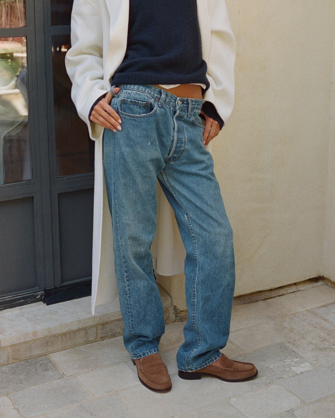 A person wearing light blue relaxed-fit denim jeans, styled with a white coat, navy top, and brown suede loafers, standing outdoors against a beige wall.