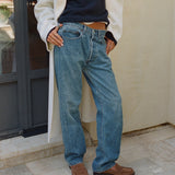 A person wearing light blue relaxed-fit denim jeans, styled with a white coat, navy top, and brown suede loafers, standing outdoors against a beige wall.