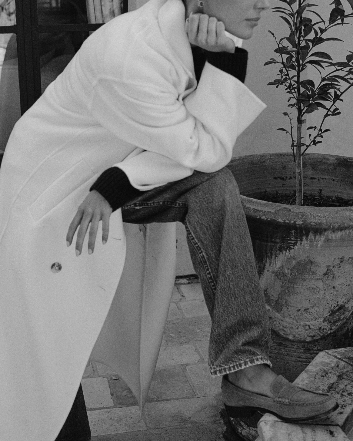 A person in a black-and-white fashion photo wearing a long white coat, dark top, wide-leg jeans, and loafers, seated beside a large potted plant.