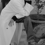 A person in a black-and-white fashion photo wearing a long white coat, dark top, wide-leg jeans, and loafers, seated beside a large potted plant.