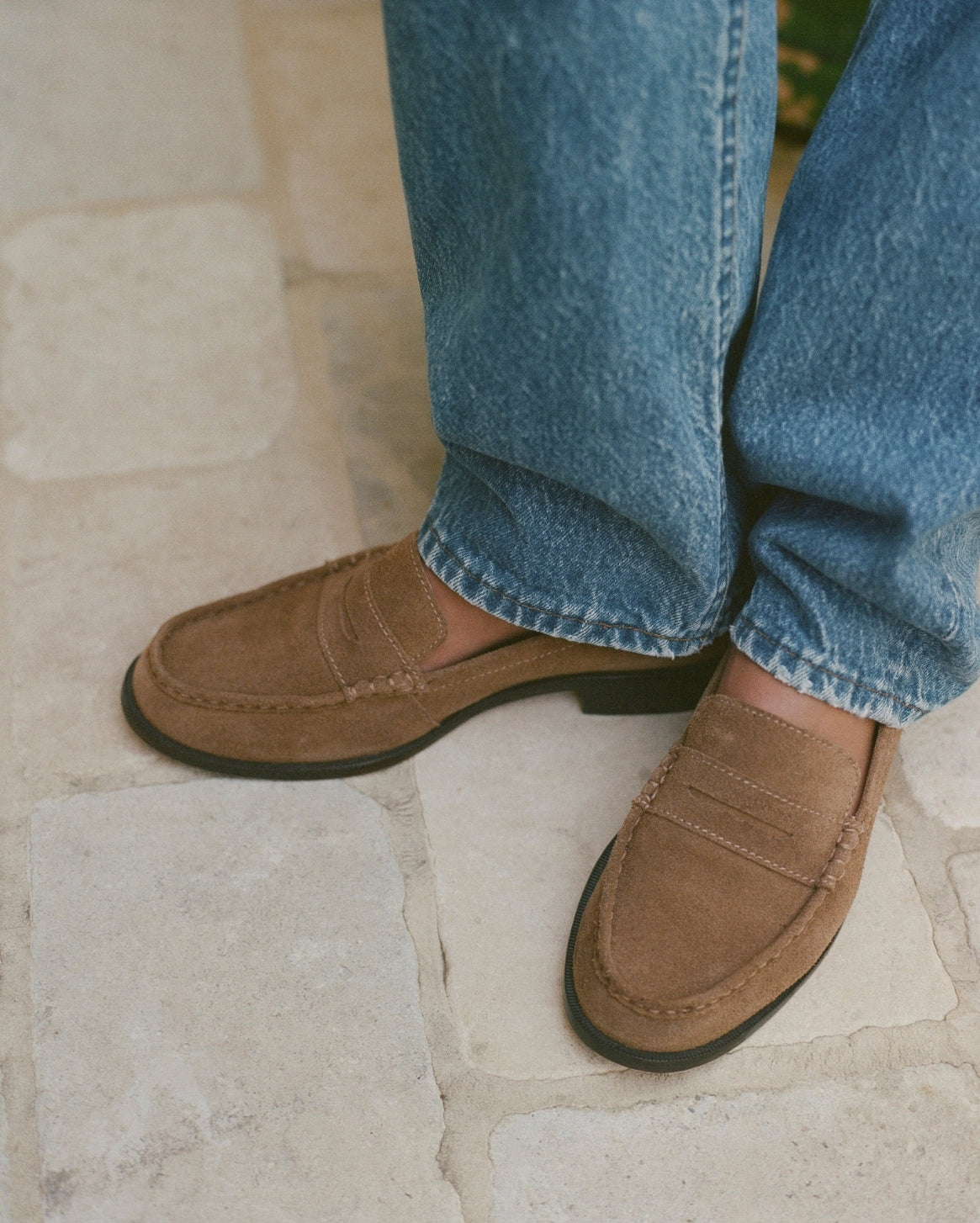 A close-up of a person wearing brown suede penny loafers with blue jeans, standing on a light beige stone tile floor.