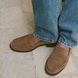 A close-up of a person wearing brown suede penny loafers with blue jeans, standing on a light beige stone tile floor.